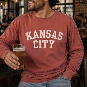 May include: A brick-red long-sleeve shirt with the text "KANSAS CITY" in white block letters. The shirt is worn with blue jeans. A glass of beer is held in the hand.