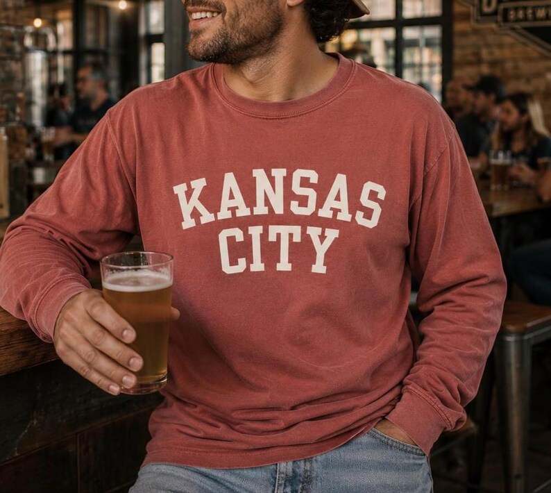May include: A brick-red long-sleeve shirt with the text "KANSAS CITY" in white block letters. The shirt is worn with blue jeans. A glass of beer is held in the hand.