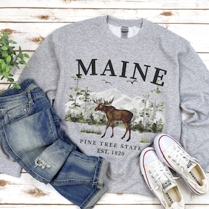 May include: Gray sweatshirt with a moose and pine trees graphic. The text "MAINE PINE TREE STATE EST. 1820" is printed on the sweatshirt.