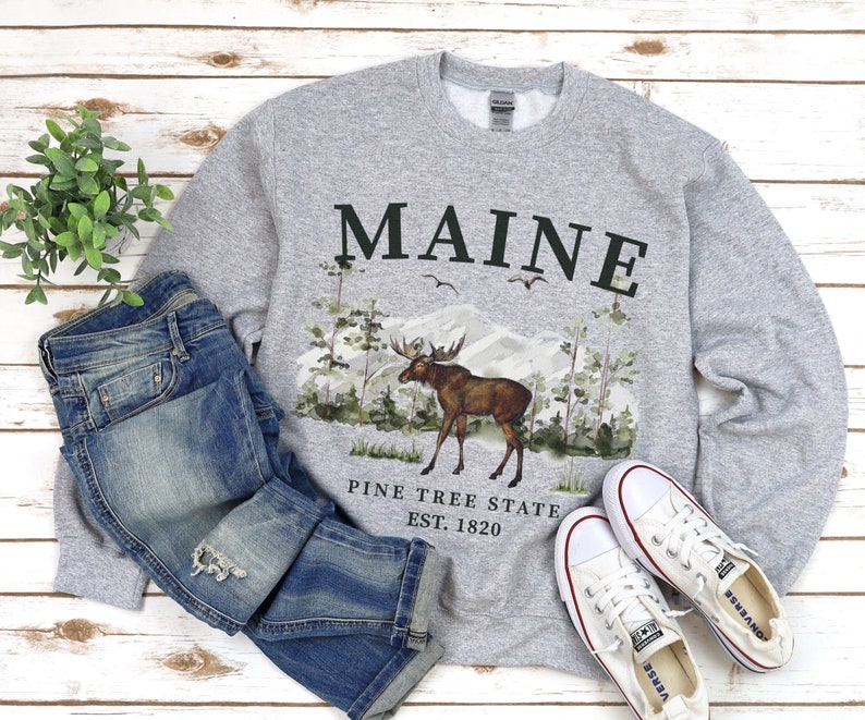 May include: Gray sweatshirt with a moose and pine trees graphic. The text "MAINE PINE TREE STATE EST. 1820" is printed on the sweatshirt.