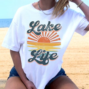 May include: White t-shirt with a retro design featuring the words "Lake Life" in a teal, distressed font. The design includes a sunset with orange and yellow hues. The shirt is a casual, comfortable style.