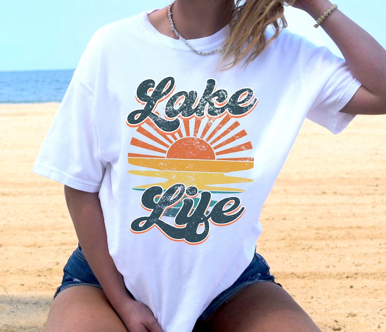 May include: White t-shirt with a retro design featuring the words "Lake Life" in a teal, distressed font. The design includes a sunset with orange and yellow hues. The shirt is a casual, comfortable style.