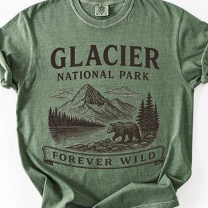 Op de afbeelding: Een saliegroen T-shirt met korte mouwen en een donkerbruine grafische print. Het ontwerp toont een berglandschap, een beer en de tekst GLACIER NATIONAL PARK en FOREVER WILD. Het shirt heeft een ronde hals en een vintage gewassen afwerking.