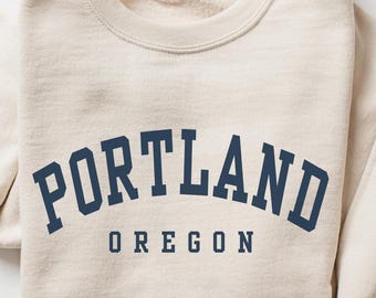 May include: A beige crewneck sweatshirt featuring the text PORTLAND in a large, dark blue, arched serif font, with the word OREGON printed in a smaller, straight, dark blue serif font directly underneath the city name.