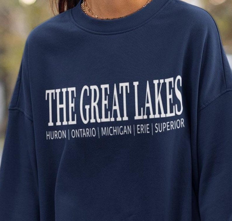 May include: A navy blue crewneck sweatshirt featuring white screen-printed text that reads THE GREAT LAKES in a large serif font, with the names HURON | ONTARIO | MICHIGAN | ERIE | SUPERIOR printed in a smaller sans-serif font directly underneath.