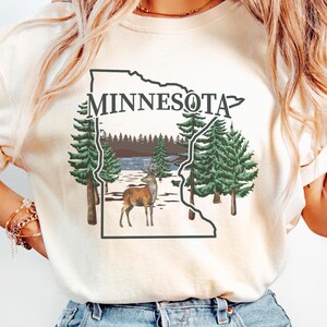 May include: A cream-colored t-shirt featuring a graphic of the state of Minnesota outlined in black. Inside the outline is a landscape with green pine trees, a brown deer, and a lake. The word MINNESOTA is printed in black above the graphic.