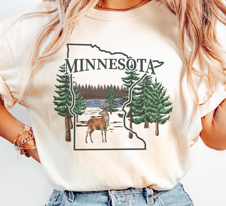 May include: A cream-colored t-shirt featuring a graphic of the state of Minnesota outlined in black. Inside the outline is a landscape with green pine trees, a brown deer, and a lake. The word MINNESOTA is printed in black above the graphic.