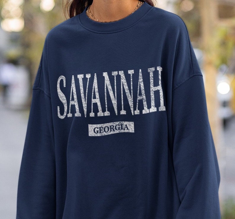 May include: Navy blue sweatshirt with the word "SAVANNAH" in distressed white lettering across the chest. Below the city name is the word "GEORGIA" in a smaller, rectangular white font. The sweatshirt has a crew neck.