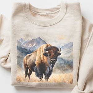 May include: A beige sweatshirt featuring a watercolor painting of a bison in a grassy field with mountains in the background. The bison is brown, black, and tan with curved horns. The artwork has a soft, natural color palette.