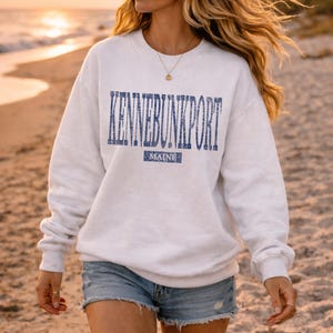 K&ouml;nnte beinhalten: Wei&szlig;es Sweatshirt mit dem Text "KENNEBUNKPORT" in Blau, darunter "MAINE". Das Sweatshirt wird mit Jeansshorts getragen. Der Hintergrund ist ein Strand bei Sonnenuntergang.