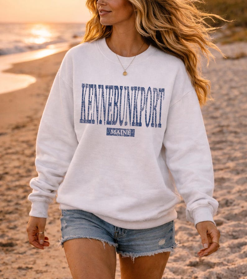 K&ouml;nnte beinhalten: Wei&szlig;es Sweatshirt mit dem Text "KENNEBUNKPORT" in Blau, darunter "MAINE". Das Sweatshirt wird mit Jeansshorts getragen. Der Hintergrund ist ein Strand bei Sonnenuntergang.