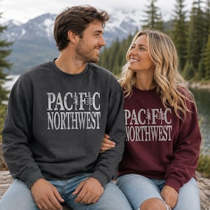 May include: A dark charcoal gray crewneck sweatshirt and a maroon crewneck sweatshirt, both featuring the white distressed text PACIFIC NORTHWEST with a pine tree graphic replacing the letter I in the word PACIFIC on the front of each garment.