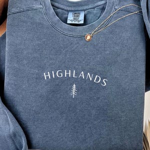 May include: A blue-grey sweatshirt with the word "HIGHLANDS" embroidered in white, and a small tree graphic. A gold necklace with a pendant rests on the sweatshirt. The tag "COMFORT COLORS" is visible on the neckline.