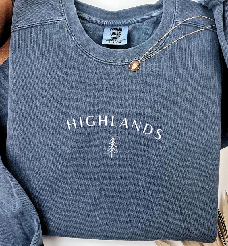 May include: A blue-grey sweatshirt with the word "HIGHLANDS" embroidered in white, and a small tree graphic. A gold necklace with a pendant rests on the sweatshirt. The tag "COMFORT COLORS" is visible on the neckline.