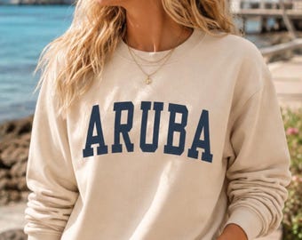 May include: A beige long-sleeve crewneck sweatshirt featuring the word ARUBA printed in large, dark navy blue block letters across the chest. The garment has a ribbed neckline, cuffs, and hem, providing a classic fit and a neutral color palette.