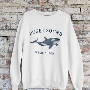 May include: A white long-sleeve crewneck sweatshirt featuring a dark blue graphic of an orca whale centered on the chest. The text "PUGET SOUND" is printed in a curved arch above the whale, and the word "WASHINGTON" is printed below the whale.