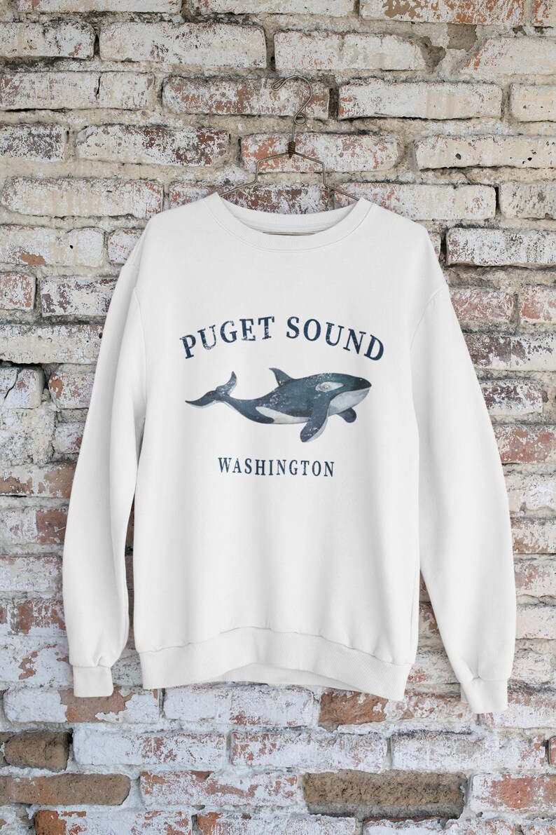 May include: A white long-sleeve crewneck sweatshirt featuring a dark blue graphic of an orca whale centered on the chest. The text "PUGET SOUND" is printed in a curved arch above the whale, and the word "WASHINGTON" is printed below the whale.