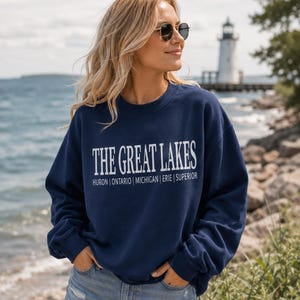 May include: A navy blue crewneck sweatshirt featuring the white text THE GREAT LAKES followed by the names HURON | ONTARIO | MICHIGAN | ERIE | SUPERIOR printed in a clean, sans-serif font across the chest area of the long-sleeved garment.