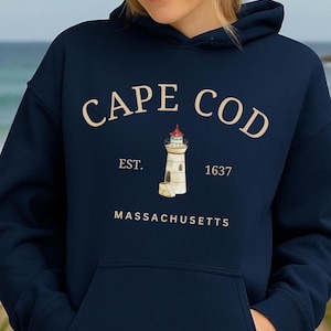 Può includere: Felpa con cappuccio blu navy con "CAPE COD" e un'illustrazione di un faro. Sotto, "EST. 1637" e "MASSACHUSETTS". La felpa ha una tasca frontale e un cappuccio con coulisse. Il design è color crema.
