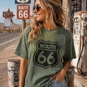 Puede incluir: Una camiseta de manga corta color verde oliva con un gr&aacute;fico negro desgastado del logotipo del escudo de la Ruta 66 en la parte delantera. La camiseta tiene cuello redondo y un corte holgado, combinada con pantalones cortos de mezclilla azul claro.