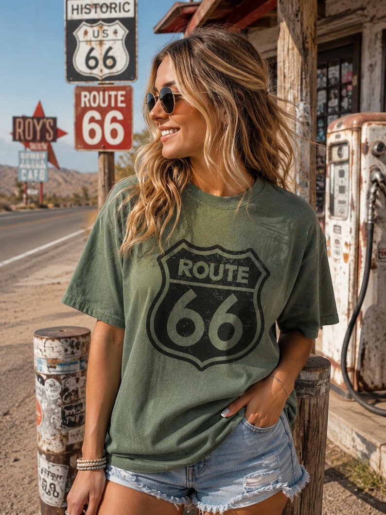 Puede incluir: Una camiseta de manga corta color verde oliva con un gr&aacute;fico negro desgastado del logotipo del escudo de la Ruta 66 en la parte delantera. La camiseta tiene cuello redondo y un corte holgado, combinada con pantalones cortos de mezclilla azul claro.