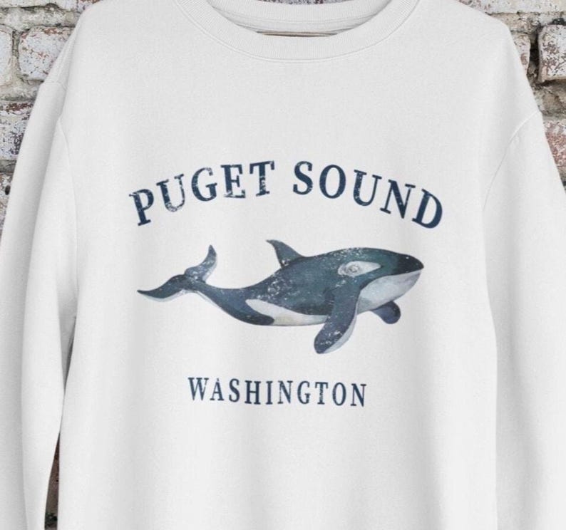 May include: A white crewneck sweatshirt featuring a dark blue graphic of an orca whale in the center. The text "PUGET SOUND" is printed in a dark blue arched font above the whale, and the word "WASHINGTON" is printed below in dark blue.