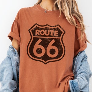 Puede incluir: Una camiseta de manga corta color &oacute;xido con un gr&aacute;fico marr&oacute;n oscuro desgastado del letrero de la carretera Route 66 centrado en el pecho. La camiseta tiene cuello redondo y se lleva debajo de una chaqueta vaquera azul claro.