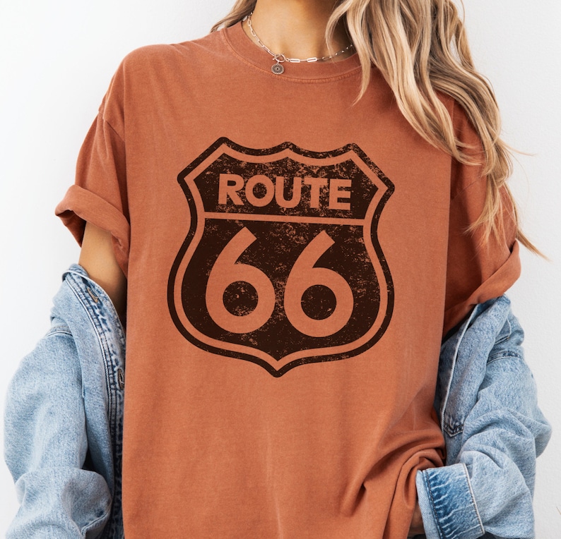 Puede incluir: Una camiseta de manga corta color &oacute;xido con un gr&aacute;fico marr&oacute;n oscuro desgastado del letrero de la carretera Route 66 centrado en el pecho. La camiseta tiene cuello redondo y se lleva debajo de una chaqueta vaquera azul claro.