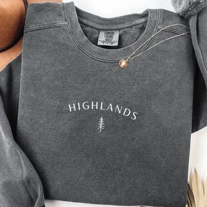 May include: A dark grey sweatshirt featuring the word "HIGHLANDS" in white script and a small tree graphic. A gold-coloured pendant necklace rests on the sweatshirt. The sweatshirt has a crew neck and long sleeves.
