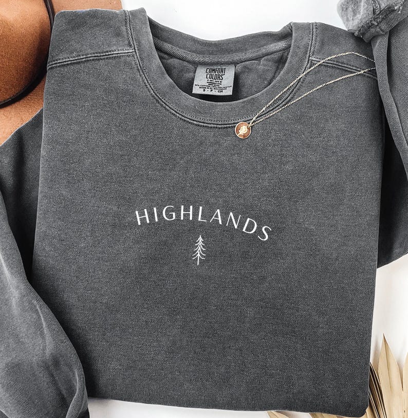 May include: A dark grey sweatshirt featuring the word "HIGHLANDS" in white script and a small tree graphic. A gold-coloured pendant necklace rests on the sweatshirt. The sweatshirt has a crew neck and long sleeves.
