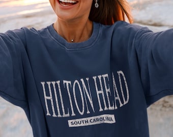 May include: A navy blue crewneck sweatshirt featuring the text HILTON HEAD in a distressed white font, with the words SOUTH CAROLINA printed in a smaller white font inside a rectangular box directly underneath the main city name.