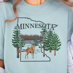 May include: A light blue t-shirt featuring a dark green outline of the state of Minnesota. Inside the outline is a graphic of a brown deer standing in a snowy forest landscape with evergreen trees and the word MINNESOTA printed in dark green.