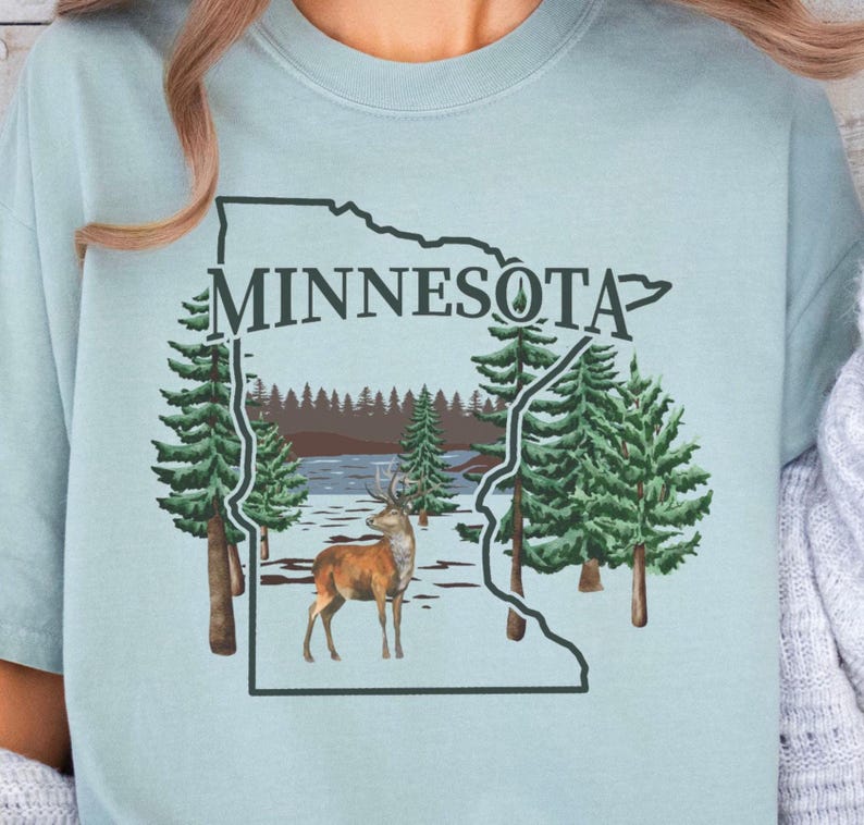 May include: A light blue t-shirt featuring a dark green outline of the state of Minnesota. Inside the outline is a graphic of a brown deer standing in a snowy forest landscape with evergreen trees and the word MINNESOTA printed in dark green.