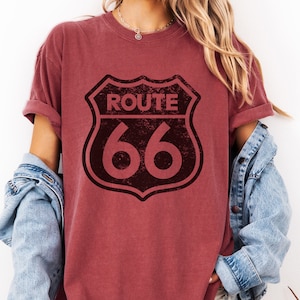 Puede incluir: Una camiseta de manga corta color granate con un gr&aacute;fico negro desgastado del escudo de la Ruta 66 en la parte delantera. La camiseta tiene cuello redondo y mangas remangadas, llevada con una chaqueta vaquera azul claro sobre los hombros.