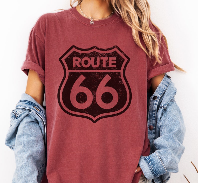 Puede incluir: Una camiseta de manga corta color granate con un gr&aacute;fico negro desgastado del escudo de la Ruta 66 en la parte delantera. La camiseta tiene cuello redondo y mangas remangadas, llevada con una chaqueta vaquera azul claro sobre los hombros.