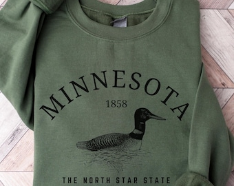 May include: An olive green crewneck sweatshirt featuring a black graphic of a loon swimming on water. The text reads MINNESOTA 1858 in an arched font above the loon, and THE NORTH STAR STATE in a straight font below the loon graphic.