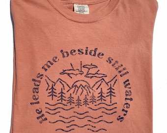 May include: A coral-colored t-shirt with a crew neck. The shirt features a navy blue circular graphic with the text "He leads me beside still waters" surrounding a mountain and tree design. The Comfort Colors tag is visible.