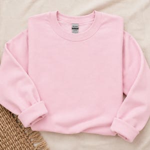 Pode incluir: Uma camisola de manga comprida e gola redonda num tom rosa claro s&oacute;lido, com punhos e bainha canelados. A pe&ccedil;a apresenta uma etiqueta da marca Gildan no interior da gola. A camisola &eacute; simples, sem quaisquer desenhos gr&aacute;ficos ou padr&otilde;es.