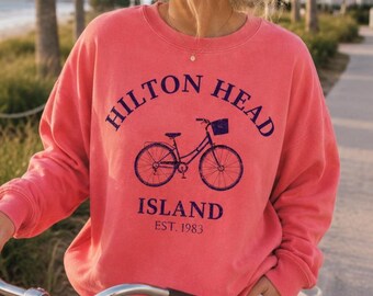 May include: A coral-colored crewneck sweatshirt featuring a dark blue graphic of a bicycle. The text reads HILTON HEAD ISLAND EST. 1983 in a dark blue font. The sweatshirt has long sleeves and a ribbed collar, cuffs, and hem.
