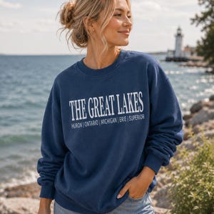 May include: A navy blue crewneck sweatshirt featuring the white text THE GREAT LAKES with the names of the lakes Huron, Ontario, Michigan, Erie, and Superior printed in a smaller font underneath the main title in a clean, sans-serif typeface.