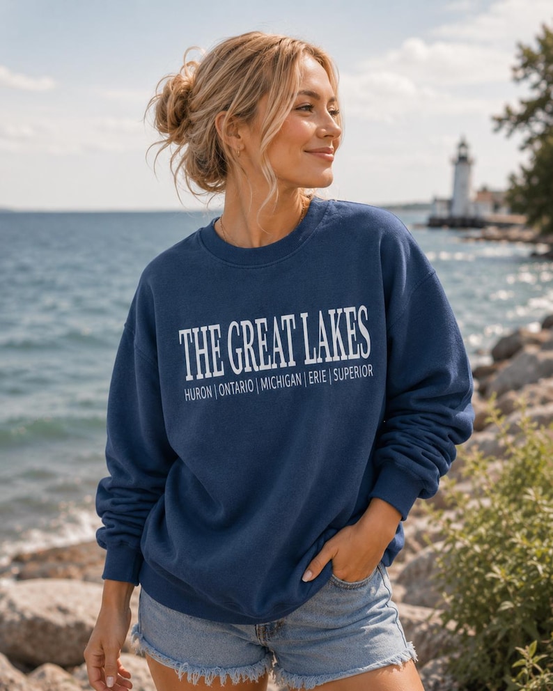 May include: A navy blue crewneck sweatshirt featuring the white text THE GREAT LAKES with the names of the lakes Huron, Ontario, Michigan, Erie, and Superior printed in a smaller font underneath the main title in a clean, sans-serif typeface.