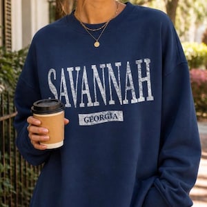 May include: Navy blue sweatshirt with the word "SAVANNAH" in large, distressed white letters. Below the word is "GEORGIA" in a smaller, rectangular white font. A person is holding a brown paper coffee cup.