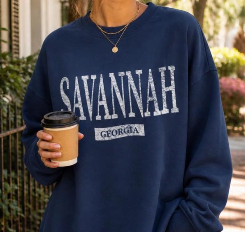 May include: Navy blue sweatshirt with the word "SAVANNAH" in large, distressed white letters. Below the word is "GEORGIA" in a smaller, rectangular white font. A person is holding a brown paper coffee cup.