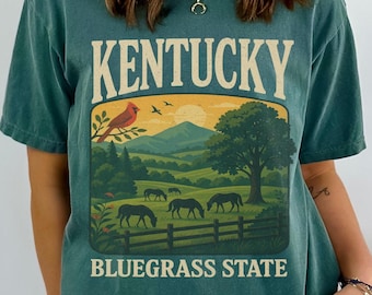 May include: A dark teal t-shirt featuring a graphic print with the text KENTUCKY and BLUEGRASS STATE. The design depicts a landscape with a cardinal bird, mountains, a large tree, and horses grazing in a fenced field under a sun.