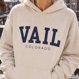 May include: A beige hooded sweatshirt featuring the word VAIL in large, dark navy blue block letters across the chest, with the word COLORADO printed in a smaller, matching navy blue font directly underneath the main text.