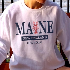 May include: A white crewneck sweatshirt featuring the dark blue text MAINE with a red lobster graphic replacing the letter I. Below this, a dark blue rectangular block contains the white text NEW ENGLAND, followed by the text EST. 1820.