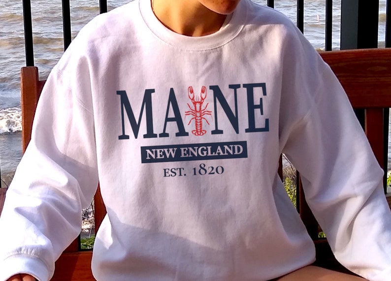 May include: A white crewneck sweatshirt featuring the dark blue text MAINE with a red lobster graphic replacing the letter I. Below this, a dark blue rectangular block contains the white text NEW ENGLAND, followed by the text EST. 1820.