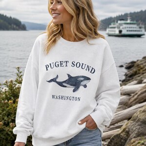 May include: A white crewneck sweatshirt featuring a dark blue graphic of an orca whale centered on the chest. The text above the whale reads "PUGET SOUND" and the text below reads "WASHINGTON" in a dark blue, capitalized serif font.