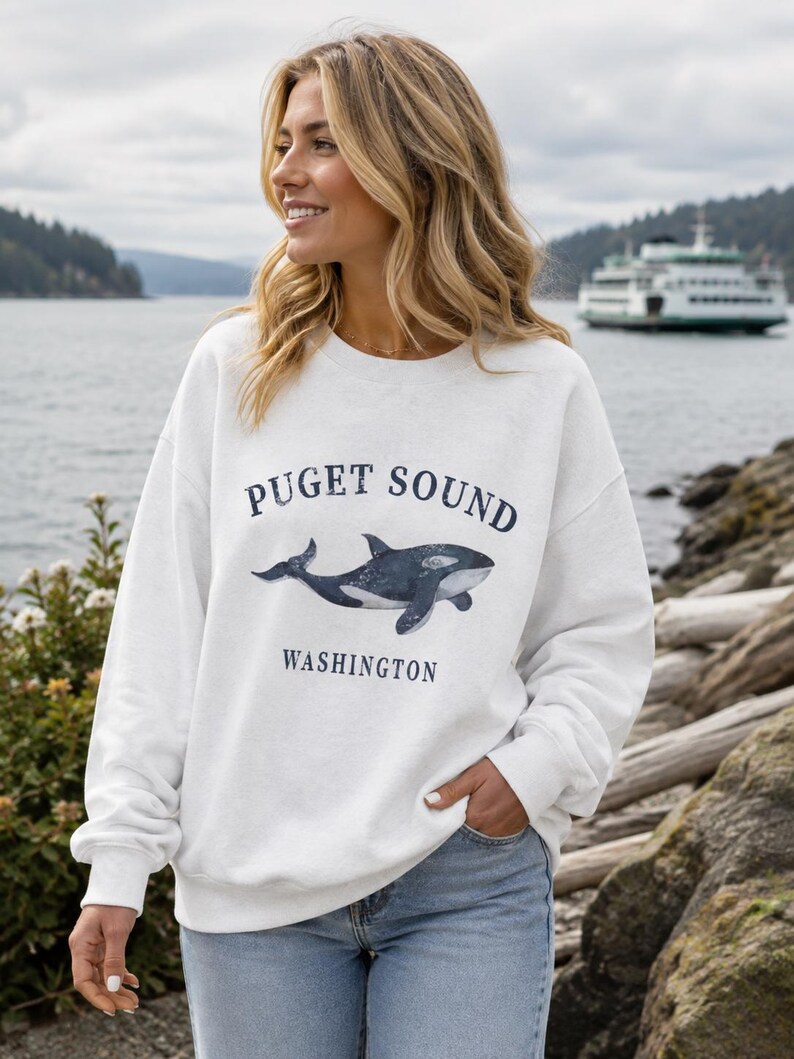 May include: A white crewneck sweatshirt featuring a dark blue graphic of an orca whale centered on the chest. The text above the whale reads "PUGET SOUND" and the text below reads "WASHINGTON" in a dark blue, capitalized serif font.