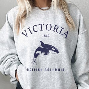 May include: A light gray crewneck sweatshirt featuring a dark blue graphic of an orca whale. The text VICTORIA 1862 is printed in a dark blue arched font above the whale, and the text BRITISH COLUMBIA is printed in dark blue below the whale.